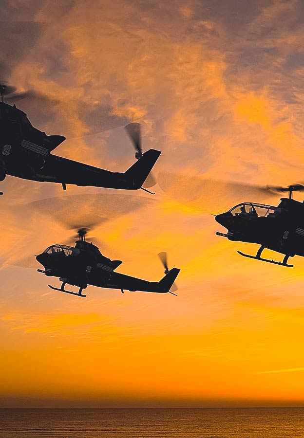 Trois hélicoptères décollent au-dessus de l’eau sur un ciel nocturne orange