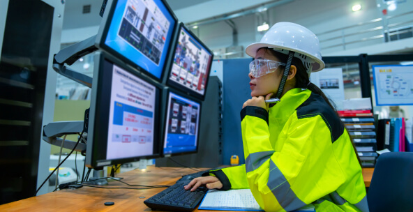 A woman in a hard hat and safety gear monitors multiple screens displaying technical data in an industrial control room.