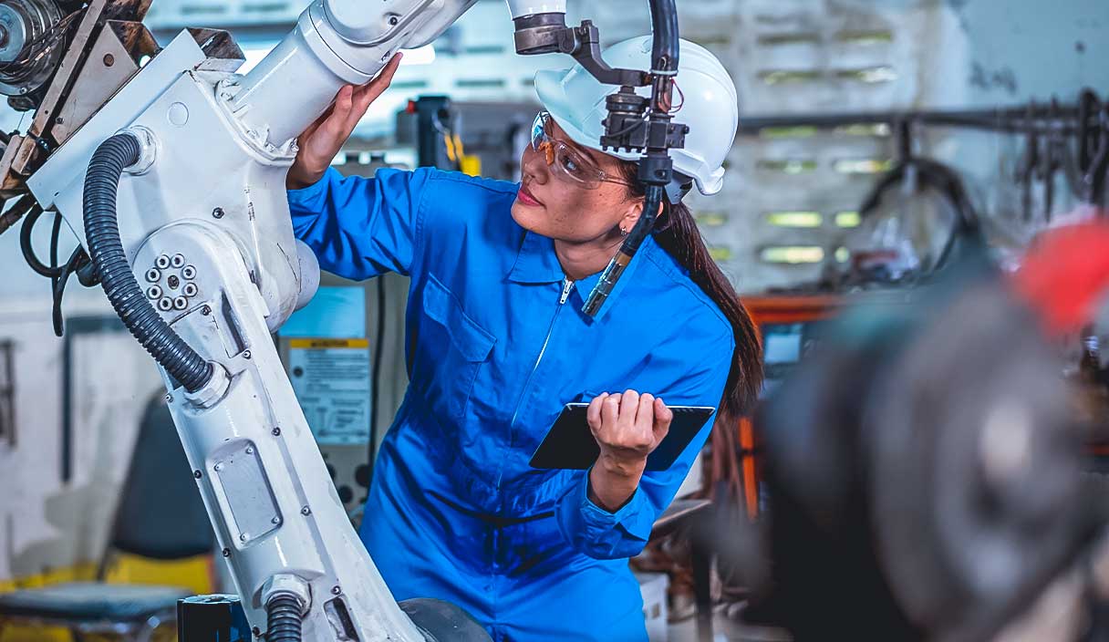Female factory worker holds tablet and checks part of robotic machine in workplace area