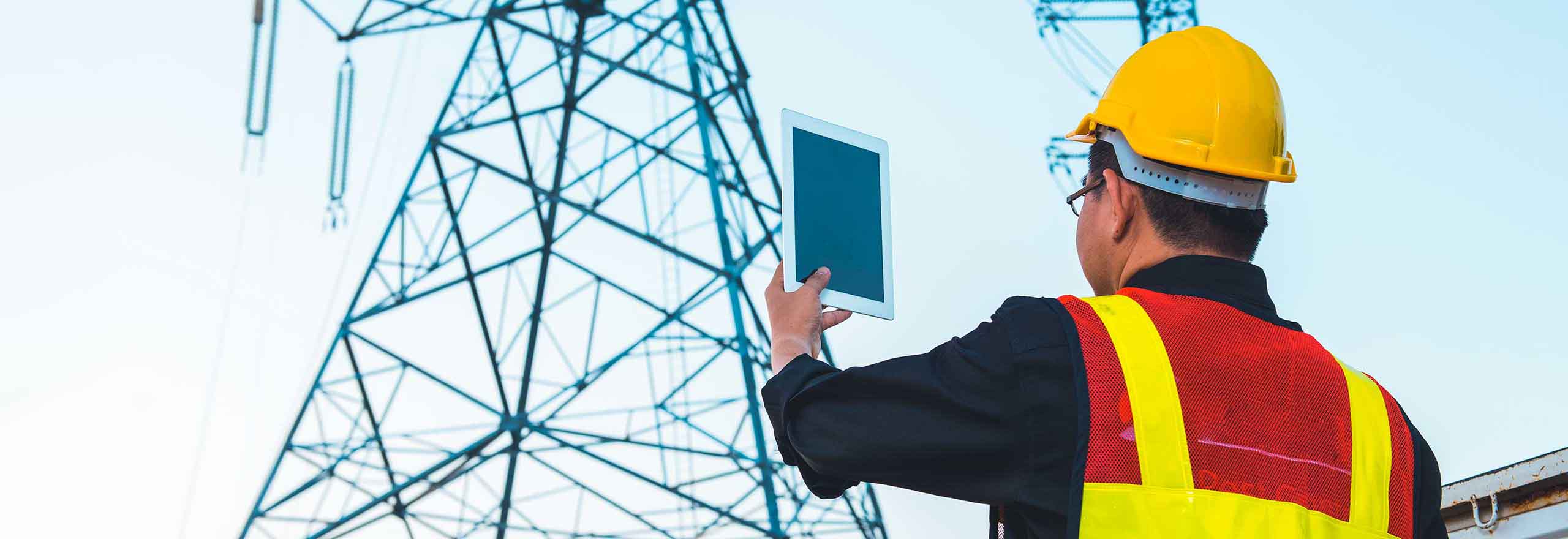 Électricien portant un casque de chantier et tenant une tablette sur un pylône d’alimentation haute tension.