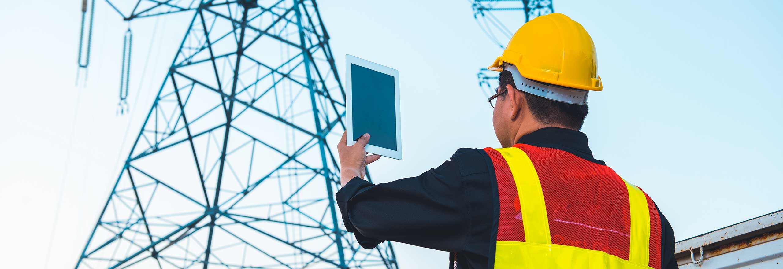 Electrical engineer in hardhat holding tablet at high-voltage power pylon.