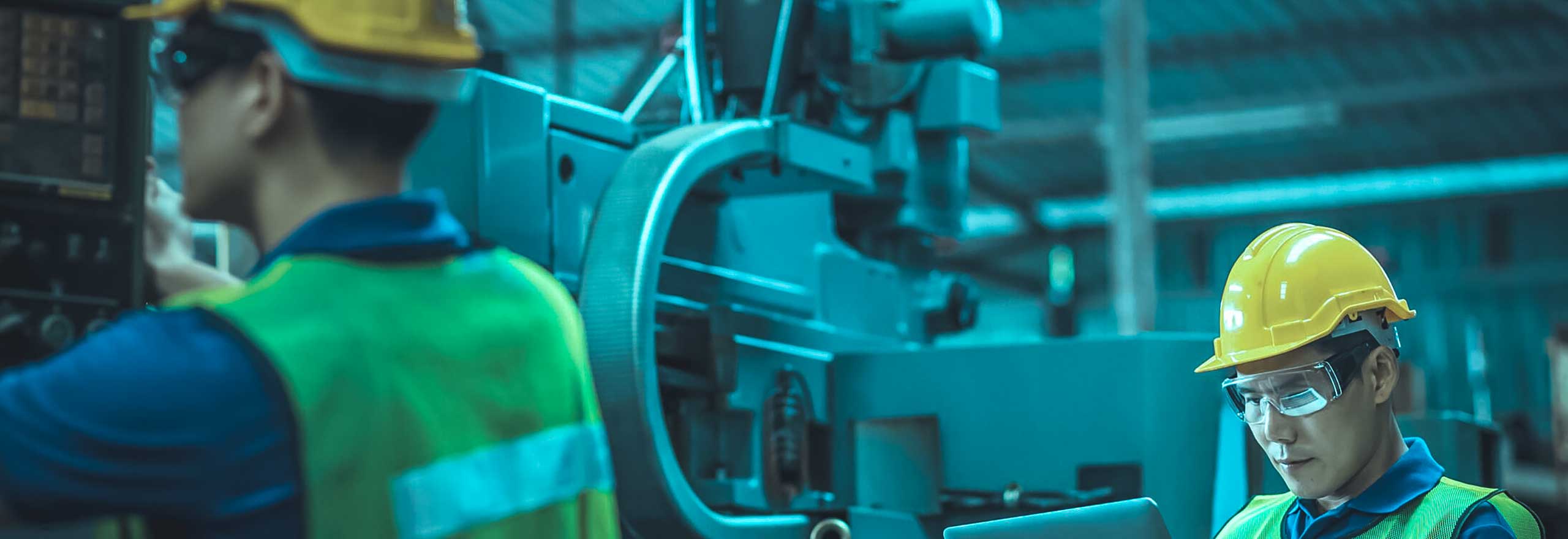 Engenheiro com capacete e um tablet analisando dados enquanto seu colega analisa a máquina em uma instalação industrial.