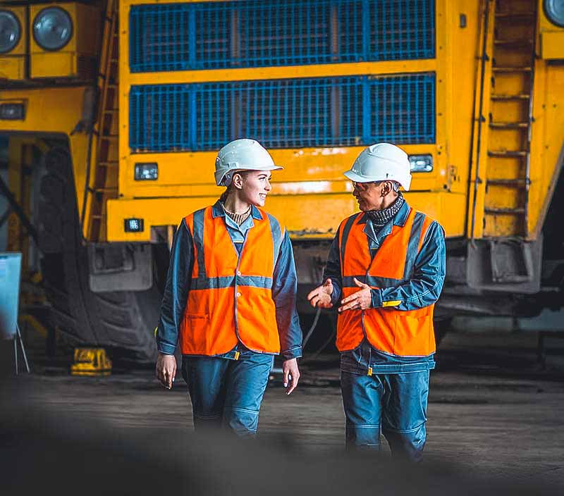 Two employees wearing hard hats and safety vests speak outdoors
