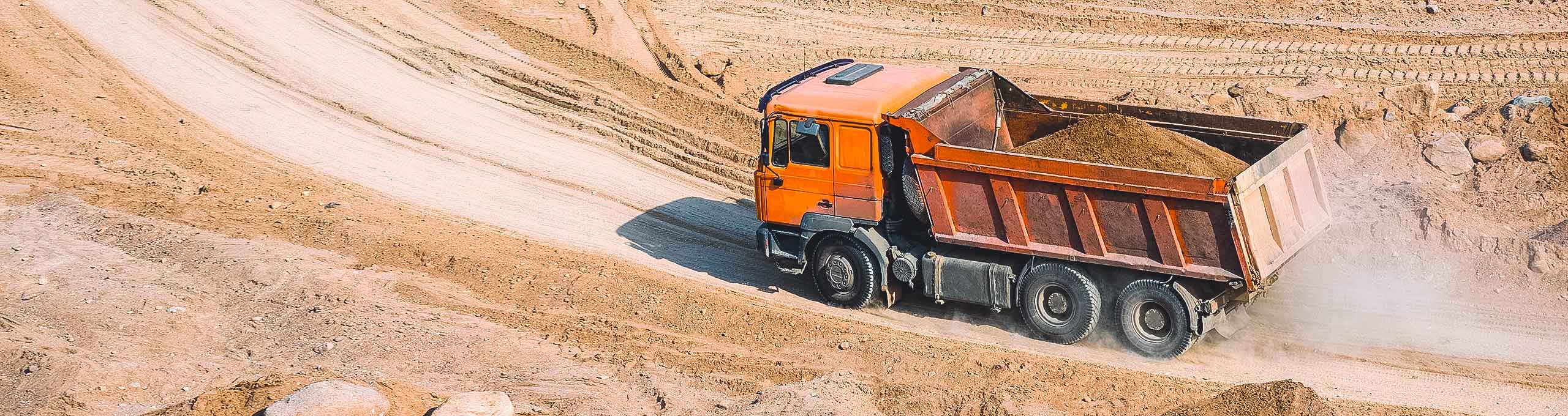 Um caminhão basculante laranja cheio de sujeira passando por uma estrada suja.