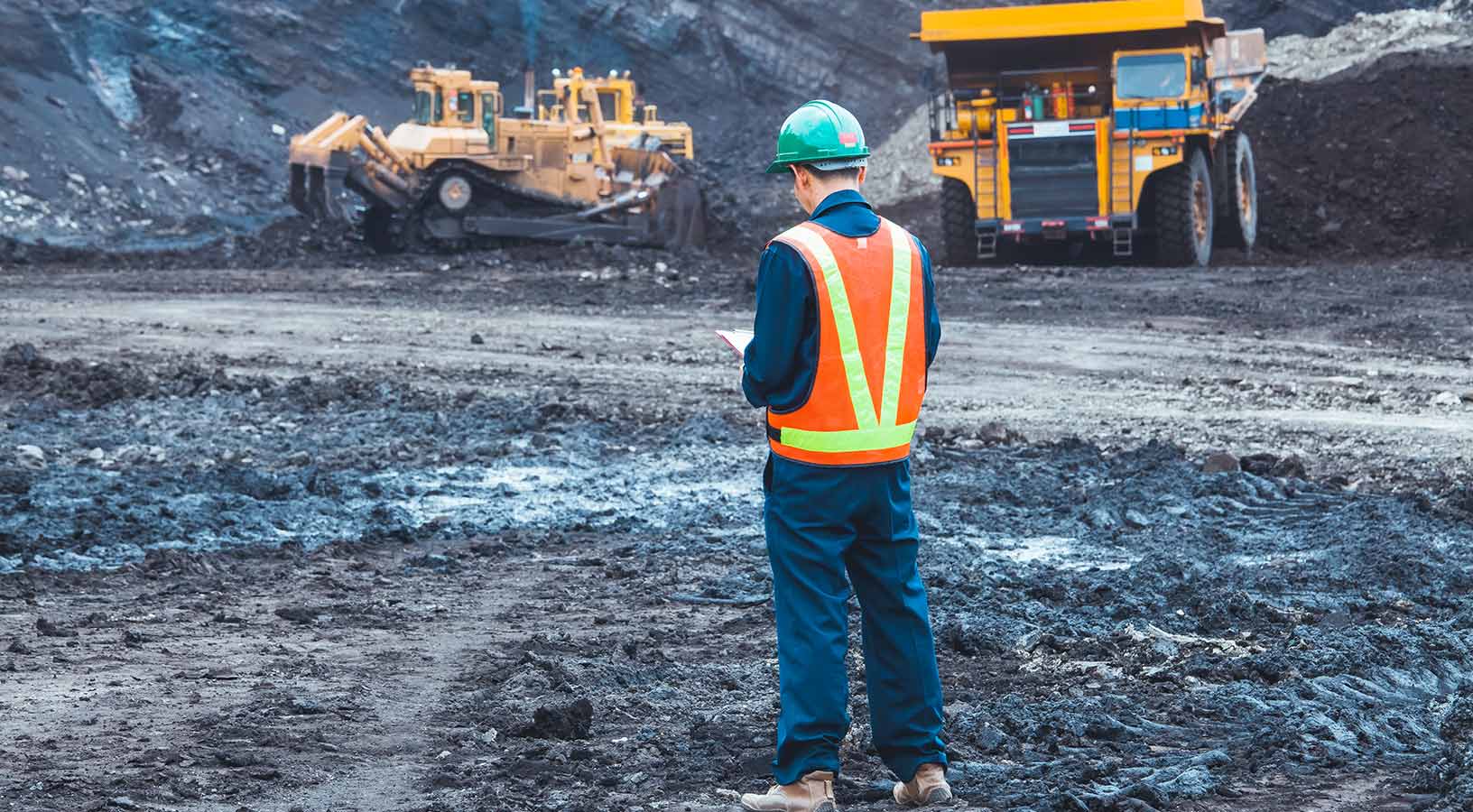 Worker in high-vis with a clipboard observing coal trucks and loaders at a mine 