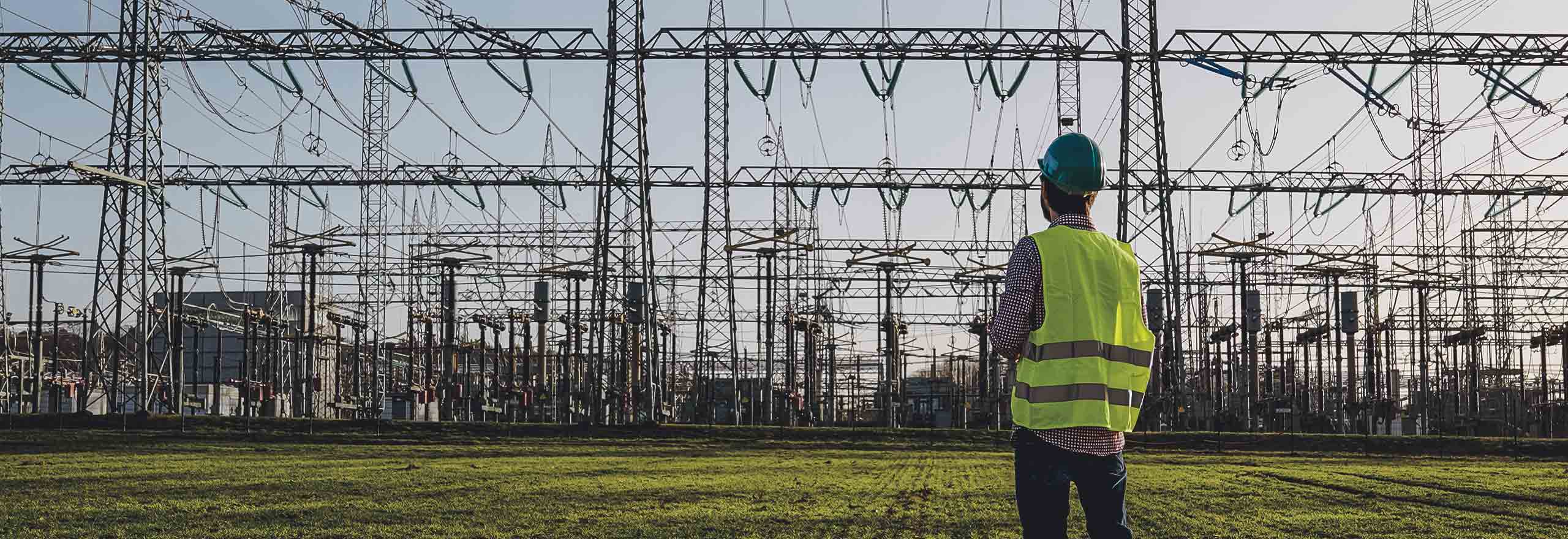 Electrical engineer wearing a helmet and safety vest working with tablet near high-voltage electrical lines power station during sunset 