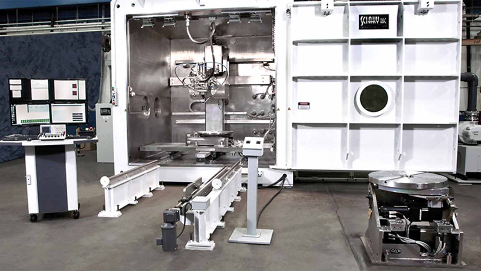 A 3D metal printer on a shop floor. In front of the printer is a  track to load parts along with a computer on a desk with four monitors