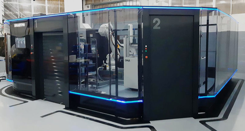 An automated cell enclosure with black and glass panelling. Inside the cell  a robotic arm can be seen loading parts onto  a shop floor coordinate measuring machine