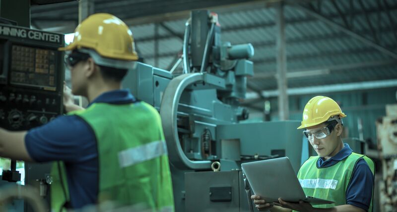 Two shopfloor workers next to machinery wearing high visibility jackets and hard hats. One of the workers is operating the machine and the other is looking at a laptop