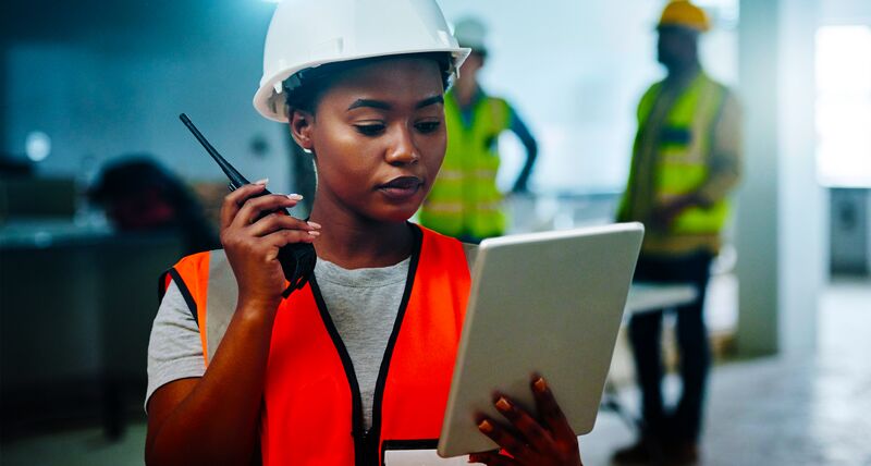 A woman wearing a hard had and high visibility jacket holding a walkie talkie and looking at a tablet device