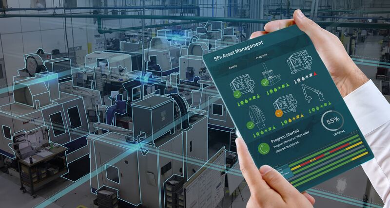 A tablet device being held in someone's hands with machine performance data on the screen. In the background the shopfloor can be seen with CNC machines 