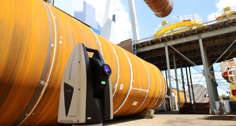 Laser tracker positioned in front of a large wind turbine component in an industrial setting, capturing measurements on a cylindrical structure outdoors.