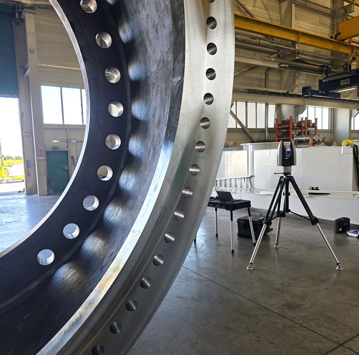 Large metal ring with circular holes in a workshop, with a tripod-mounted laser tracker nearby.