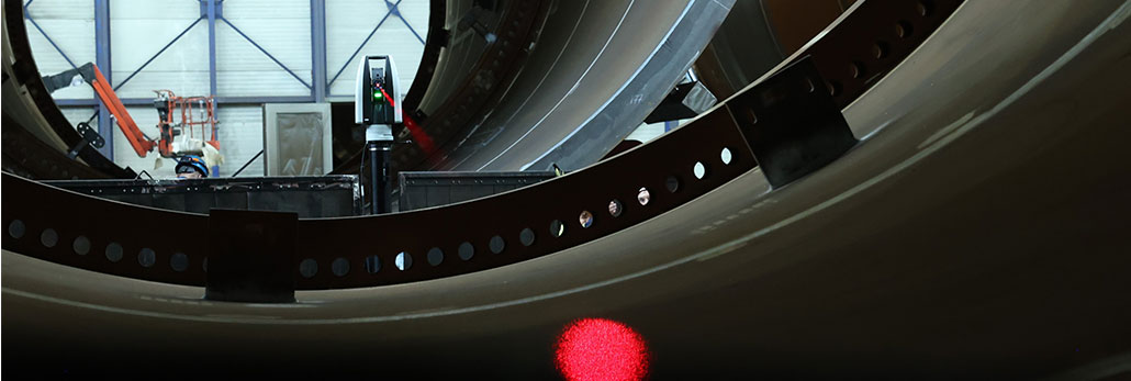 A large industrial metal ring structure inside a manufacturing facility, with a laser tracker positioned near the inner edge.