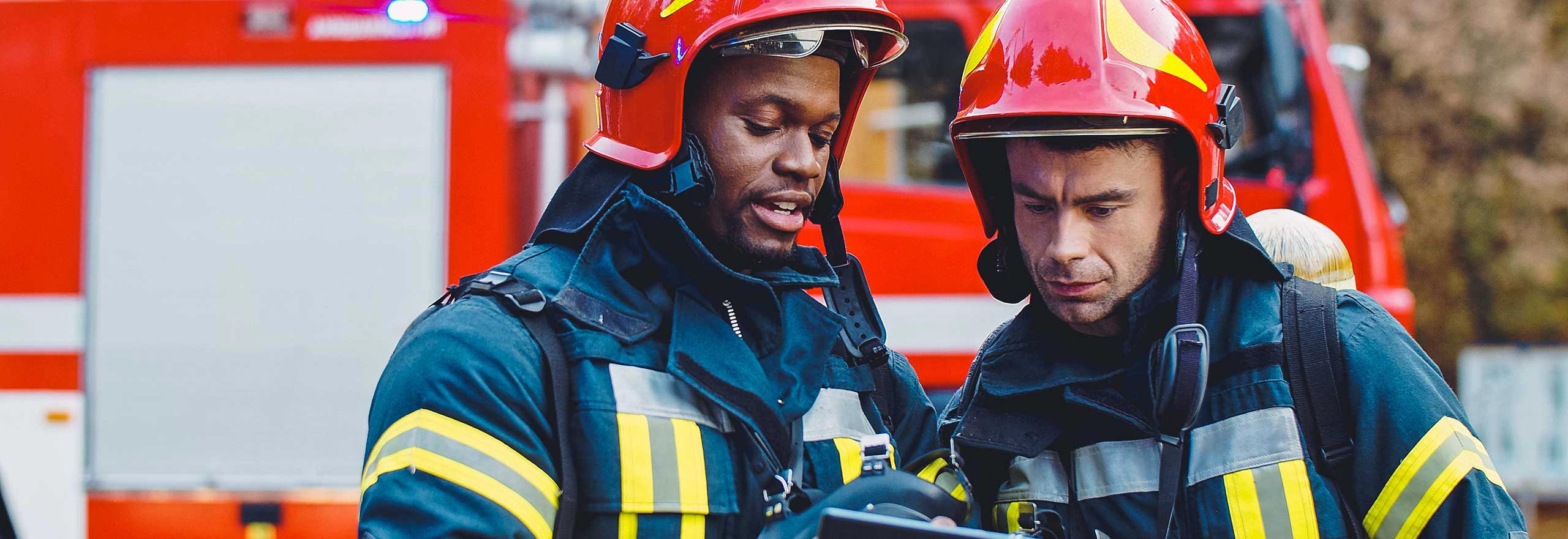 Retrato de dois bombeiros em operação de combate a incêndio, bombeiro com roupas de proteção e capacete utilizando um tablet em ação de combate.
