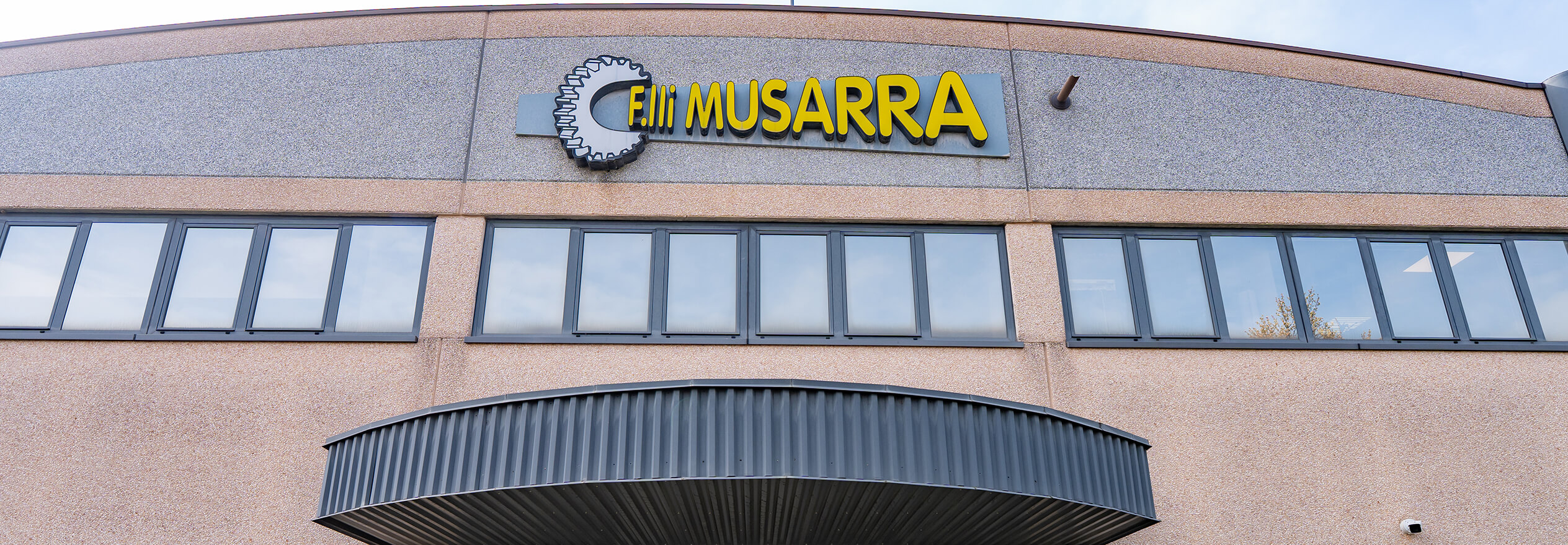 F.lli Musarra factory entrance with yellow gear logo above glass doors on a beige industrial building