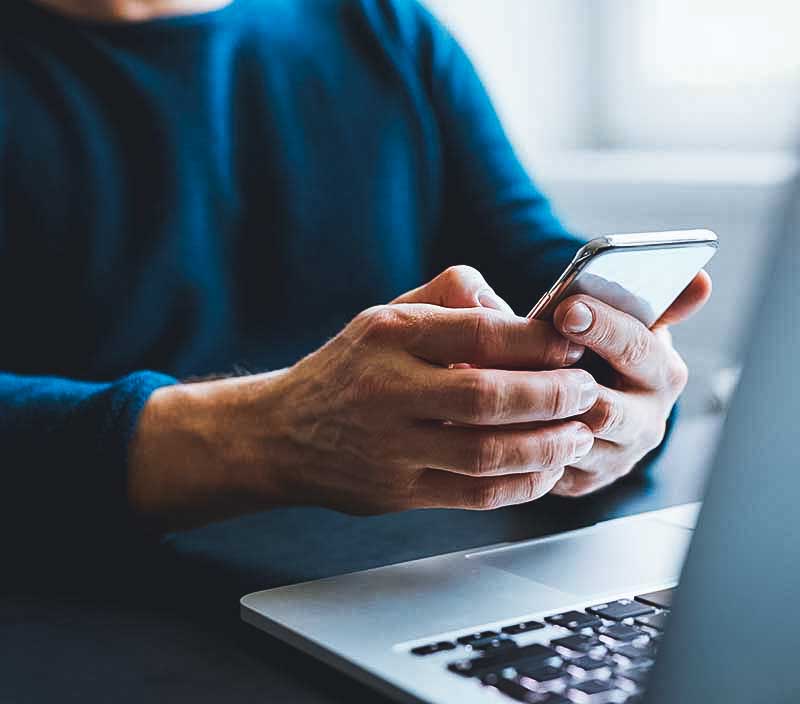 Uma pessoa segurando e olhando para um celular enquanto está sentado em frente a um laptop