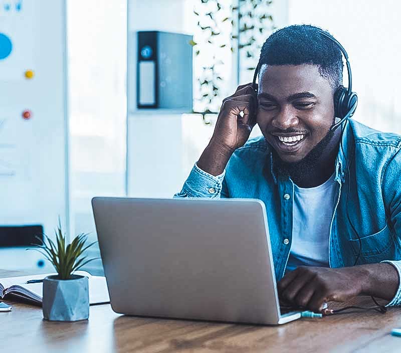 A smiling man in an office wearing headphones looks at his laptop