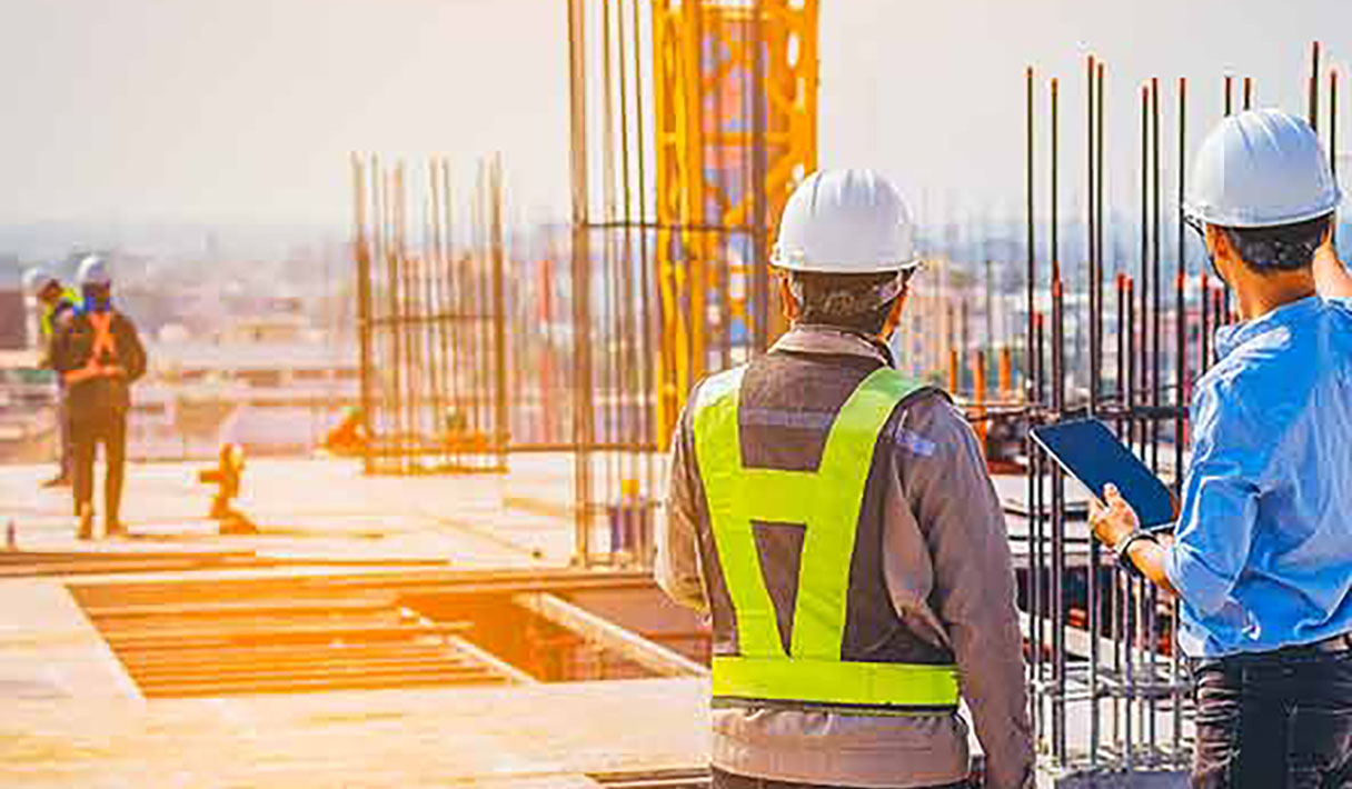 Un empleado de construcción visualizando datos en una tableta y de pie frente a un proyecto de construcción y otros empleados